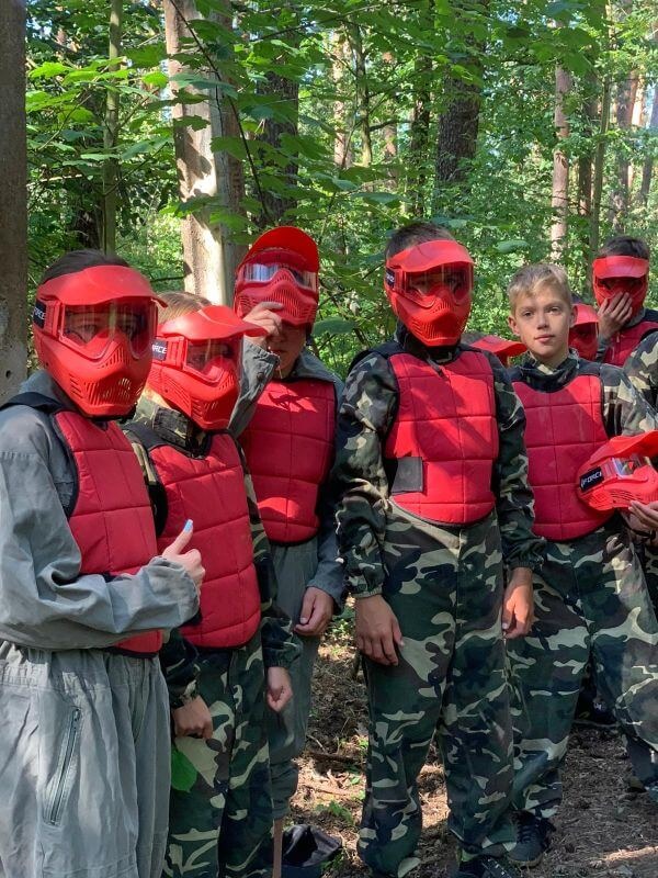 Grupa dzieci w lesie, ubrana w czerwone maski i kamizelki ochronne, w strojach moro, przygotowana do gry w paintball, jeden z uczestników pokazuje kciuk w górę.