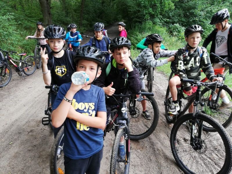Grupa dzieci w kaskach na rowerach na leśnej ścieżce, jeden chłopiec pije wodę z butelki.