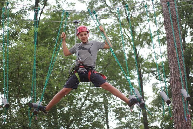 Chłopiec w czerwonym kasku, z szeroko rozstawionymi nogami, pokonuje przeszkodę w parku linowym, trzymając się zielonych lin.