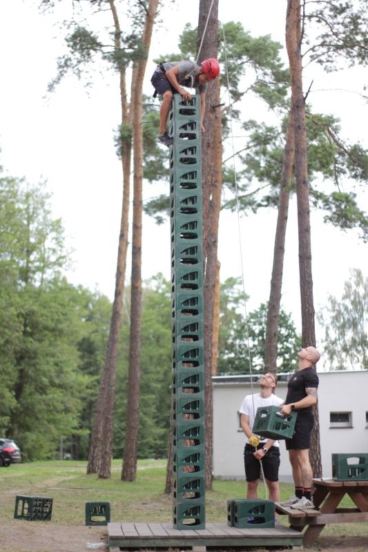 Uczestnik w czerwonym kasku wspina się na wieżę z plastikowych skrzynek, obserwowany przez dwóch mężczyzn na ziemi, teren leśny.