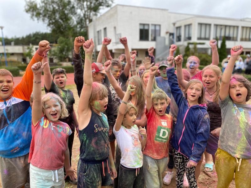 Grupa roześmianych dzieci z uniesionymi rękami, pokrytych kolorowymi proszkami podczas zabawy na świeżym powietrzu, na tle budynku.