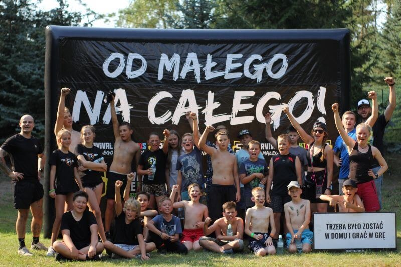 Grupa dzieci i dorosłych świętuje sukces przed czarnym banerem z napisem 'OD MAŁEGO NA CAŁEGO!' i tabliczką 'TRZEBA BYŁO ZOSTAĆ W DOMU I GRAĆ W GRĘ', wszyscy trzymają ręce w górze w geście triumfu.