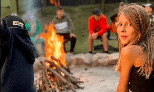 Uśmiechnięta dziewczynka z warkoczykami w tle ogniska i siedzących osób na piasku.