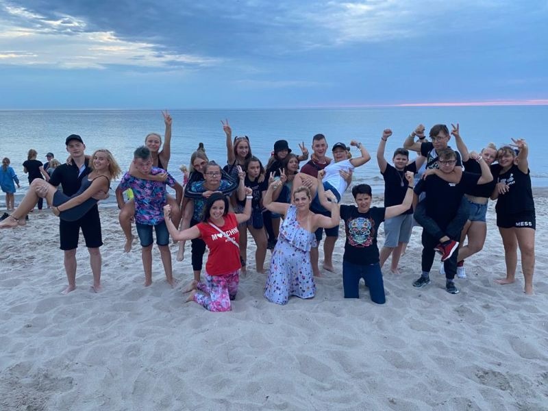 Grupa młodzieży pozuje na piaszczystej plaży nad morzem, niektórzy podnoszą ręce w geście radości, inni niosą się nawzajem, w tle zachodzące słońce i spokojna woda.