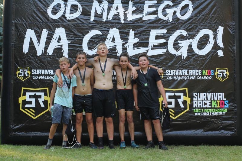 Grupa pięciu chłopców z medalami po biegu z przeszkodami Survival Race Kids, stojących na tle baneru z hasłem 'Od małego na całego!'