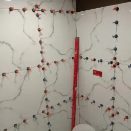 Białe płytki imitujące marmur na ścianie łazienki, wyrównane za pomocą klipsów i klinów w kolorach pomarańczowym i niebieskim, pionowy poziom w rogu, widoczna puszka elektryczna.