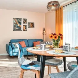 Jasne wnętrze pokoju dziennego z niebieską sofą, okrągłym drewnianym stołem z czarnymi nogami, krzesłami w patchworkowe wzory i pomarańczowymi akcentami w postaci zasłon i poduszek.