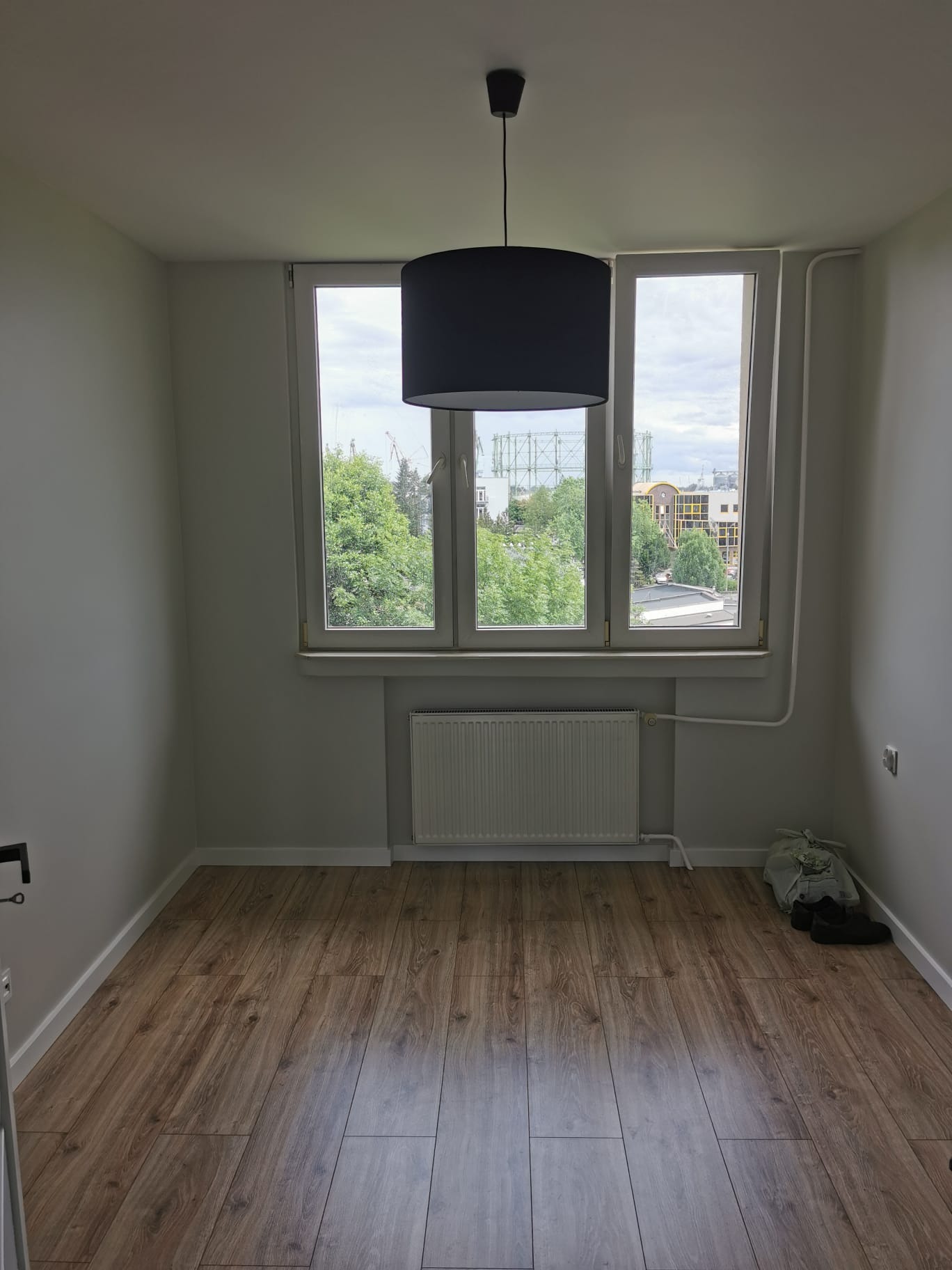Pusty pokój po położeniu paneli podłogowych z widokiem na miasto przez okno, z zamontowanym grzejnikiem i wiszącą lampą.