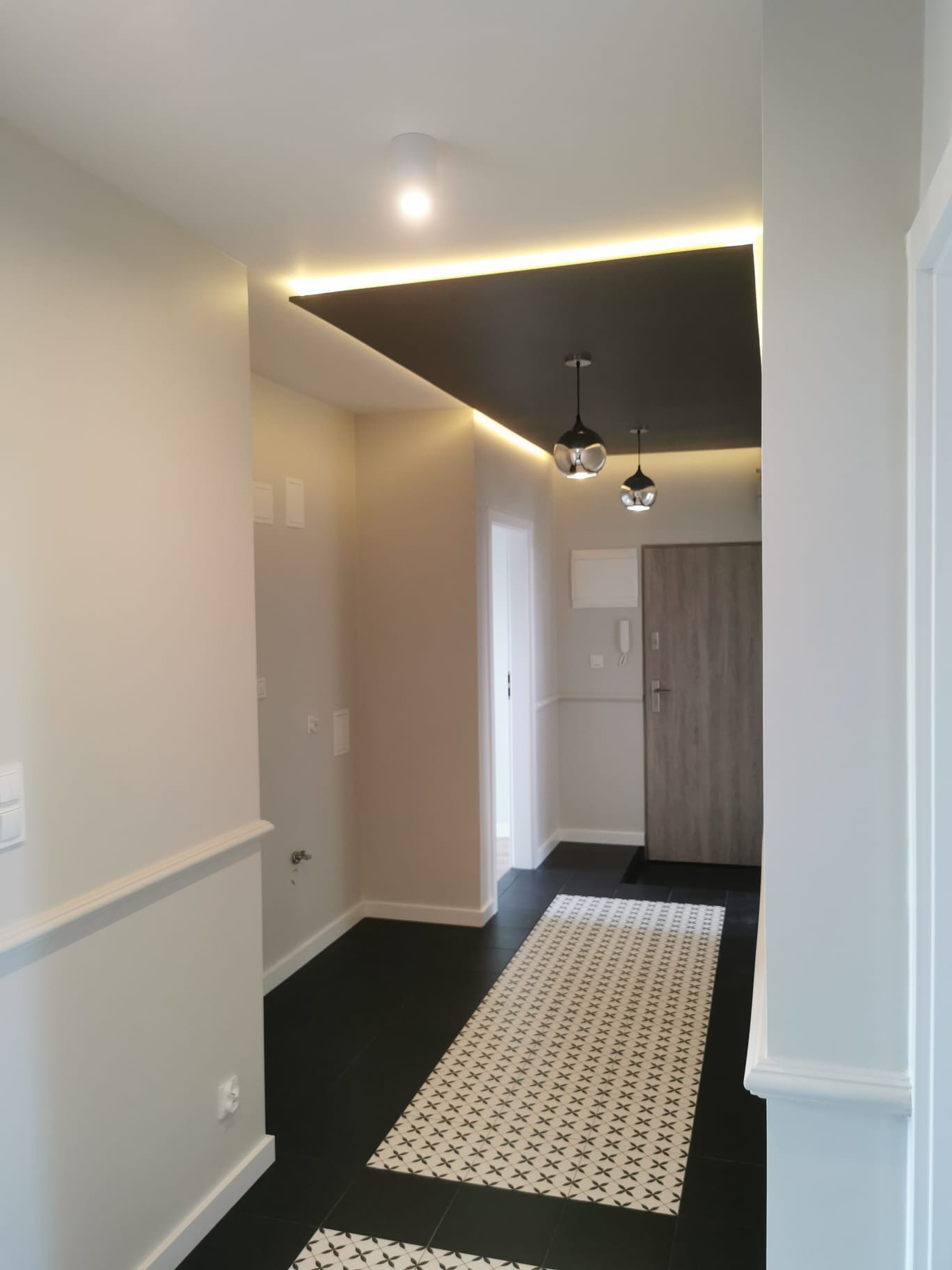 Nowoczesny przedpokój z czarną podwieszaną częścią sufitu z oświetleniem LED, czarną podłogą z białym wzorem, oraz wiszącymi lampami w kształcie kuli.