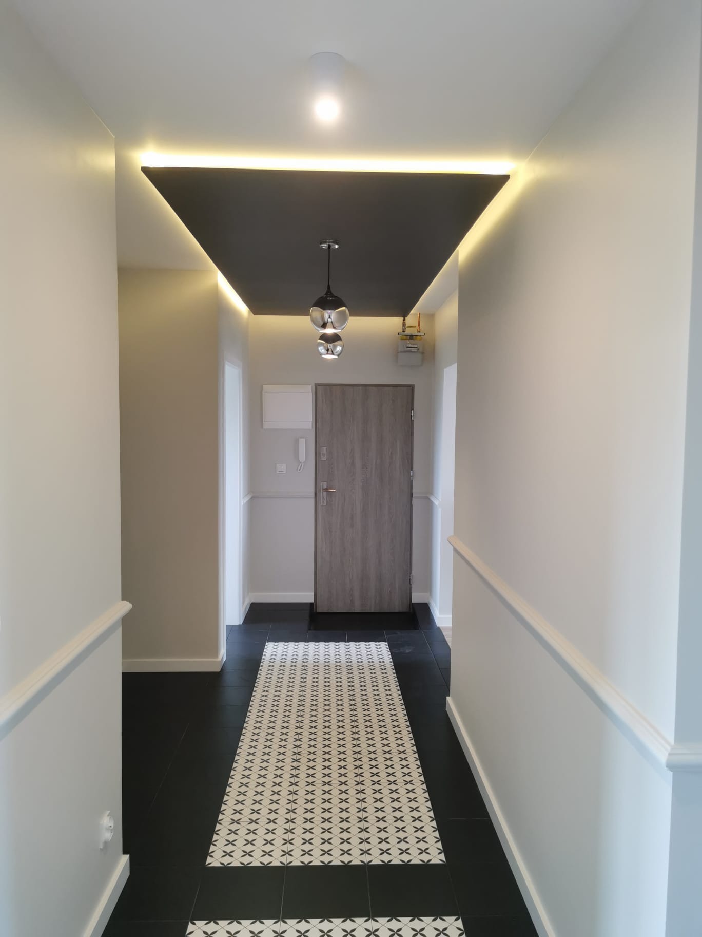 Korytarz po renowacji z czarno-białą podłogą, ciemnym sufitem podwieszanym z oświetleniem LED i nowoczesną lampą wiszącą.