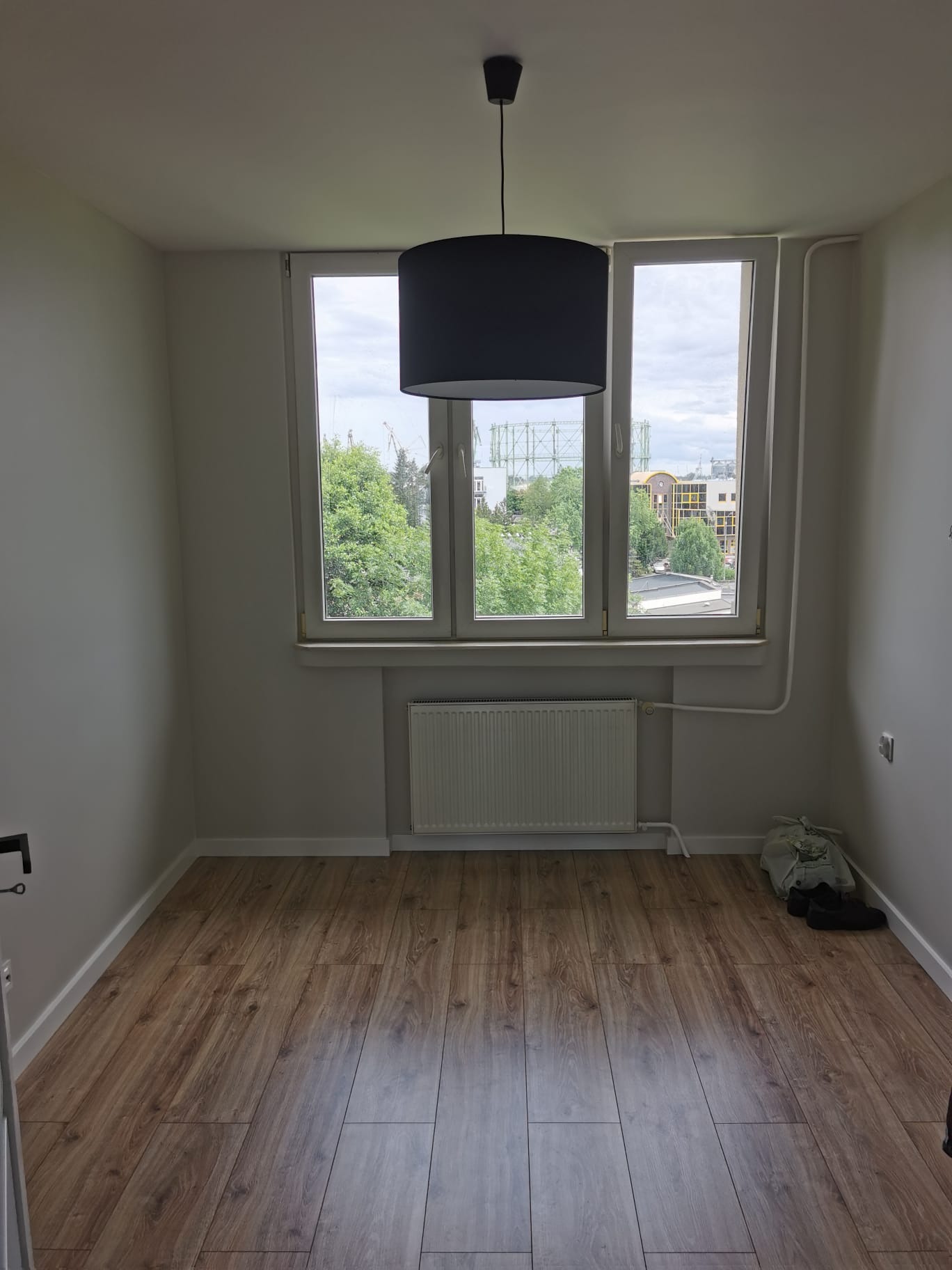 Pusty, jasny pokój po remoncie z panelami podłogowymi imitującymi drewno, białymi ścianami, grzejnikiem pod oknem i wiszącą lampą z czarnym abażurem.