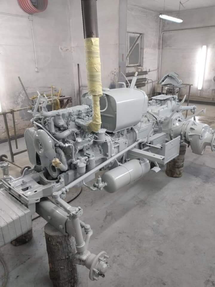 Częściowo zdemontowany traktor, świeżo pomalowany na szaro, stojący na drewnianych klockach w warsztacie, z rurą wydechową owiniętą taśmą malarską.