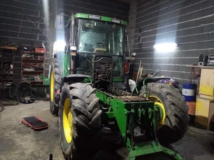 Zielony traktor John Deere w trakcie naprawy w warsztacie, widoczne opony z żółtymi felgami i brakująca maska, na podłodze leży podkładka warsztatowa, ściany z szarego betonu.