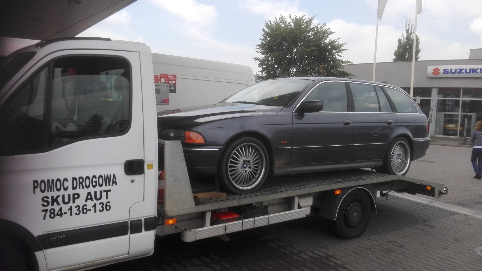 Laweta z ciemnoszarym BMW kombi na platformie, w tle salon Suzuki.