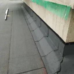 Szary dach płaski z papy termozgrzewalnej, widoczny fragment ściany budynku z zabezpieczeniem siatką, obok leży poziomica.