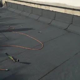 Nowo położona, czarna papa termozgrzewalna na dachu budynku w Krzywdzie, widoczne narzędzia dekarskie i przewód gazowy.