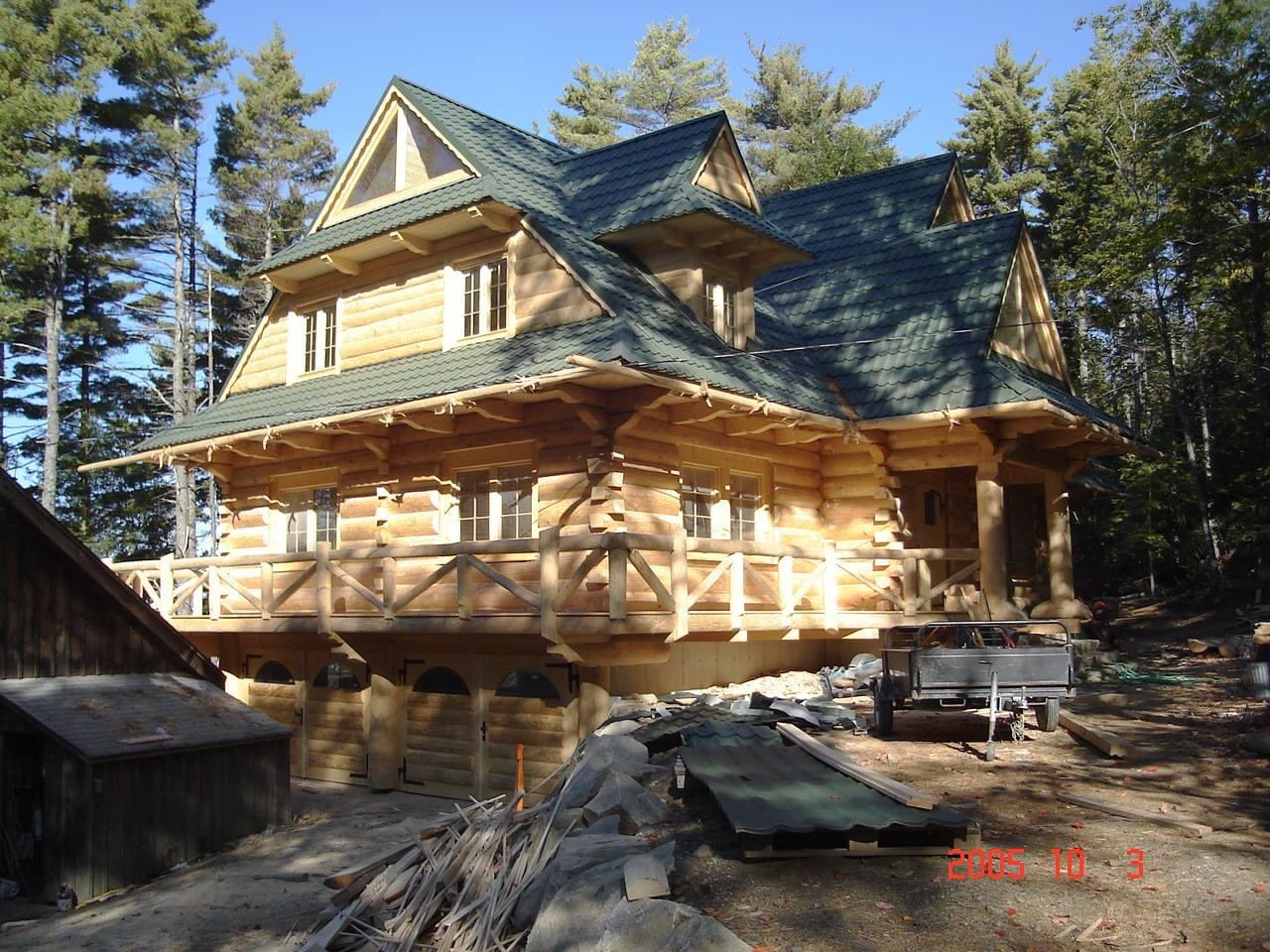 Dwupiętrowy dom z bali z zielonym dachem, balkonem i garażem pod spodem w otoczeniu drzew. Widoczne materiały budowlane i przyczepka.