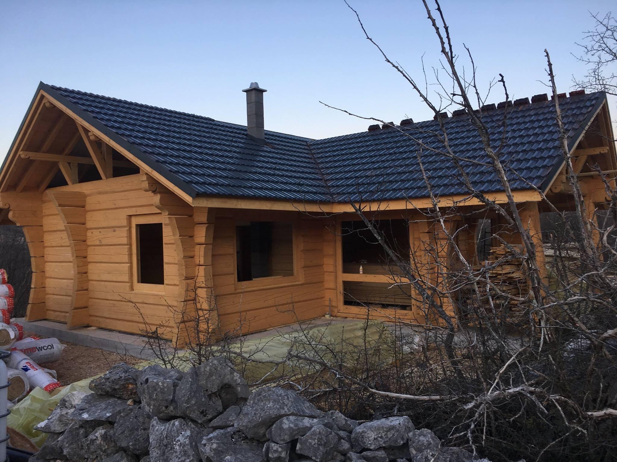 Surowa bryła drewnianego domu z ciemnym dachem z dachówki, widoczna konstrukcja więźby dachowej, stos materiałów budowlanych i kamienny murek w tle.