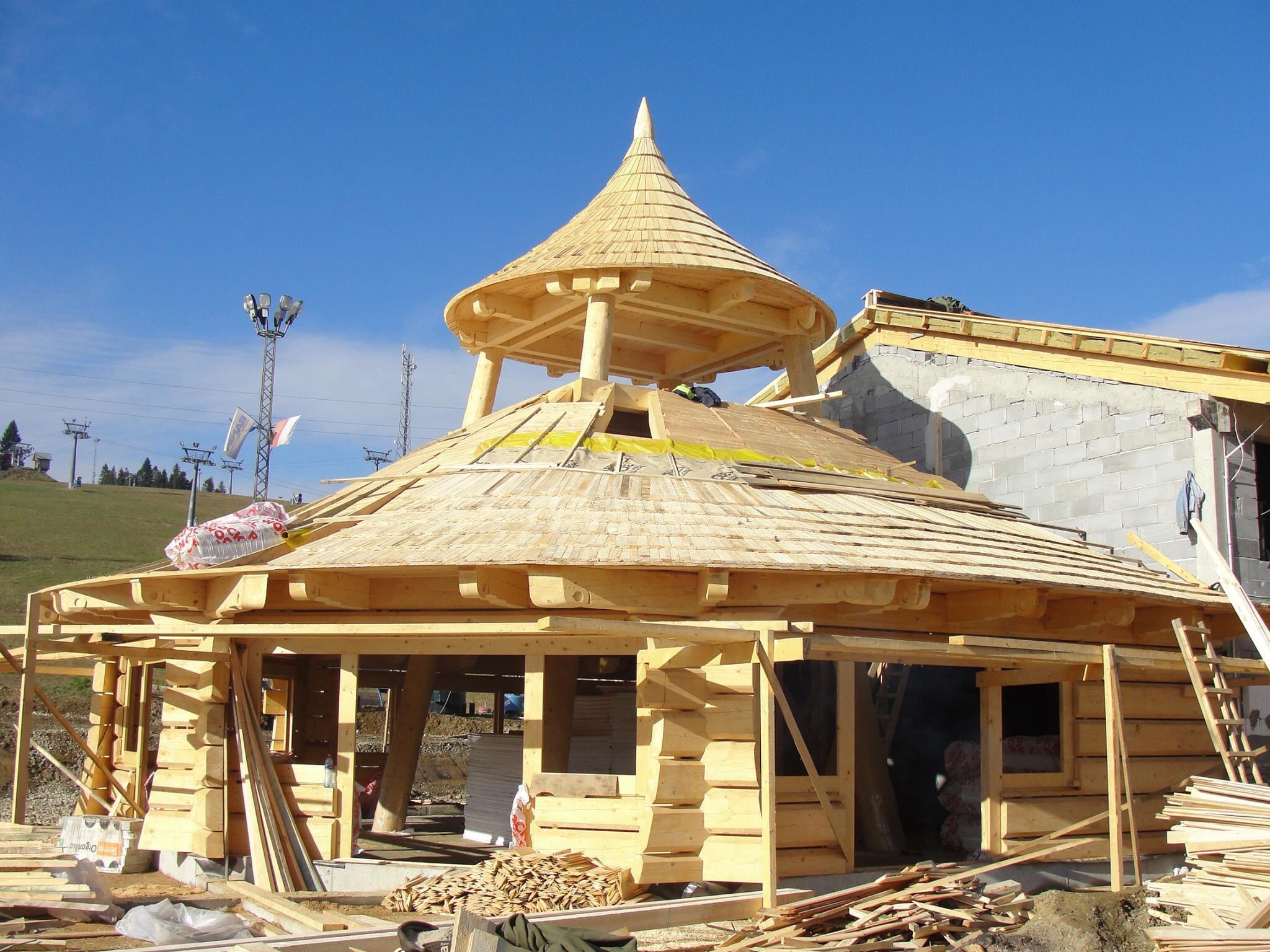 Konstrukcja drewniana z dachem z gontu w trakcie budowy, widoczne elementy rusztowania i materiały budowlane, w tle wzgórza i wyciąg narciarski.