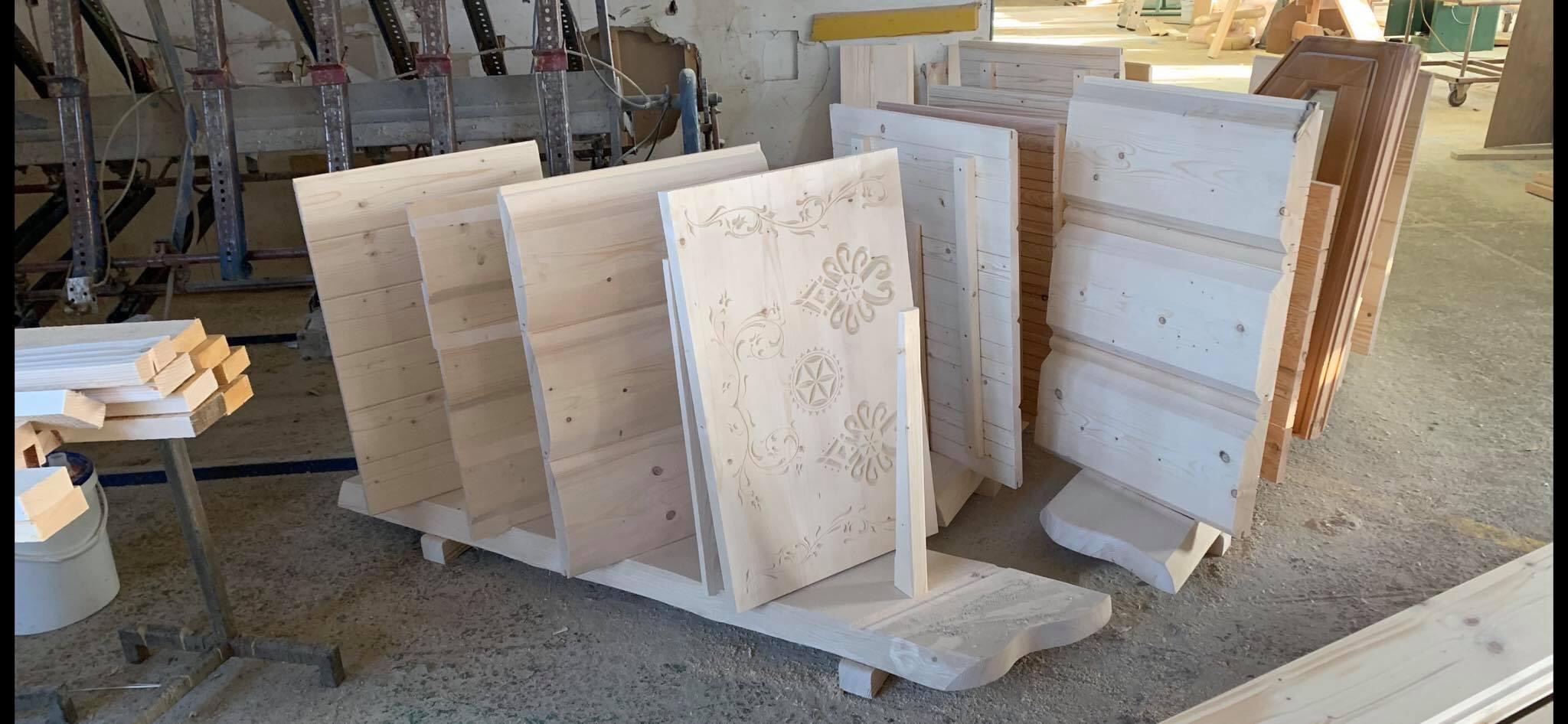 Elementy drewniane ścian z bali, niektóre z dekoracyjnymi wzorami, ustawione w warsztacie przed montażem.