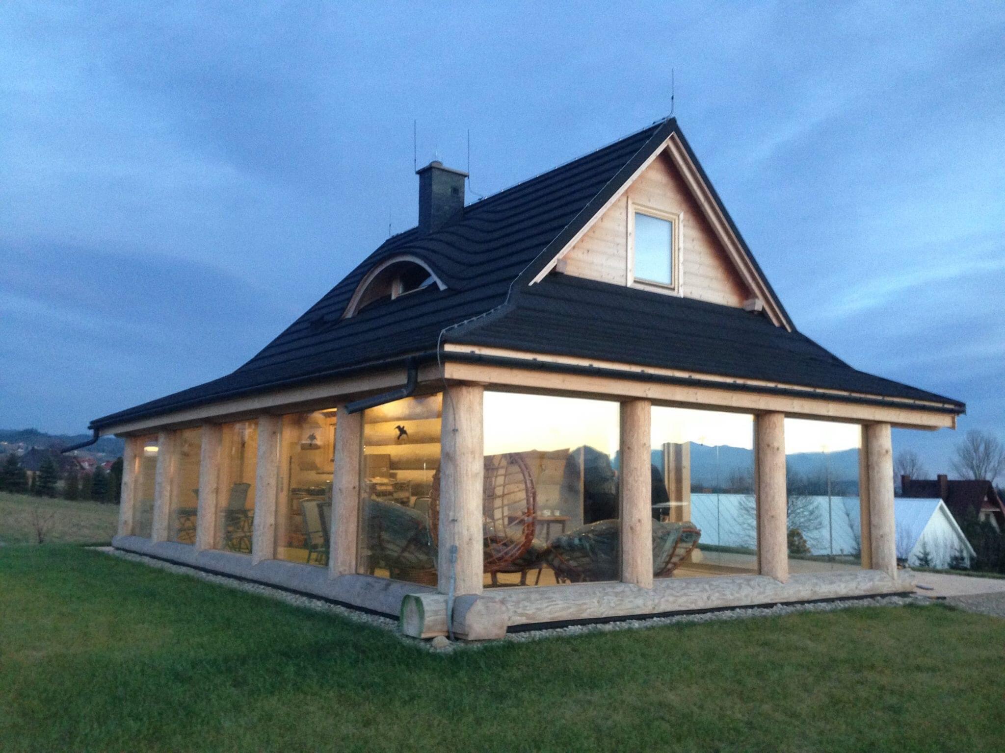 Nowoczesny dom z drewnianych bali z przeszkloną werandą i czarnym dachem, widok z zewnątrz na tle zachodzącego słońca.