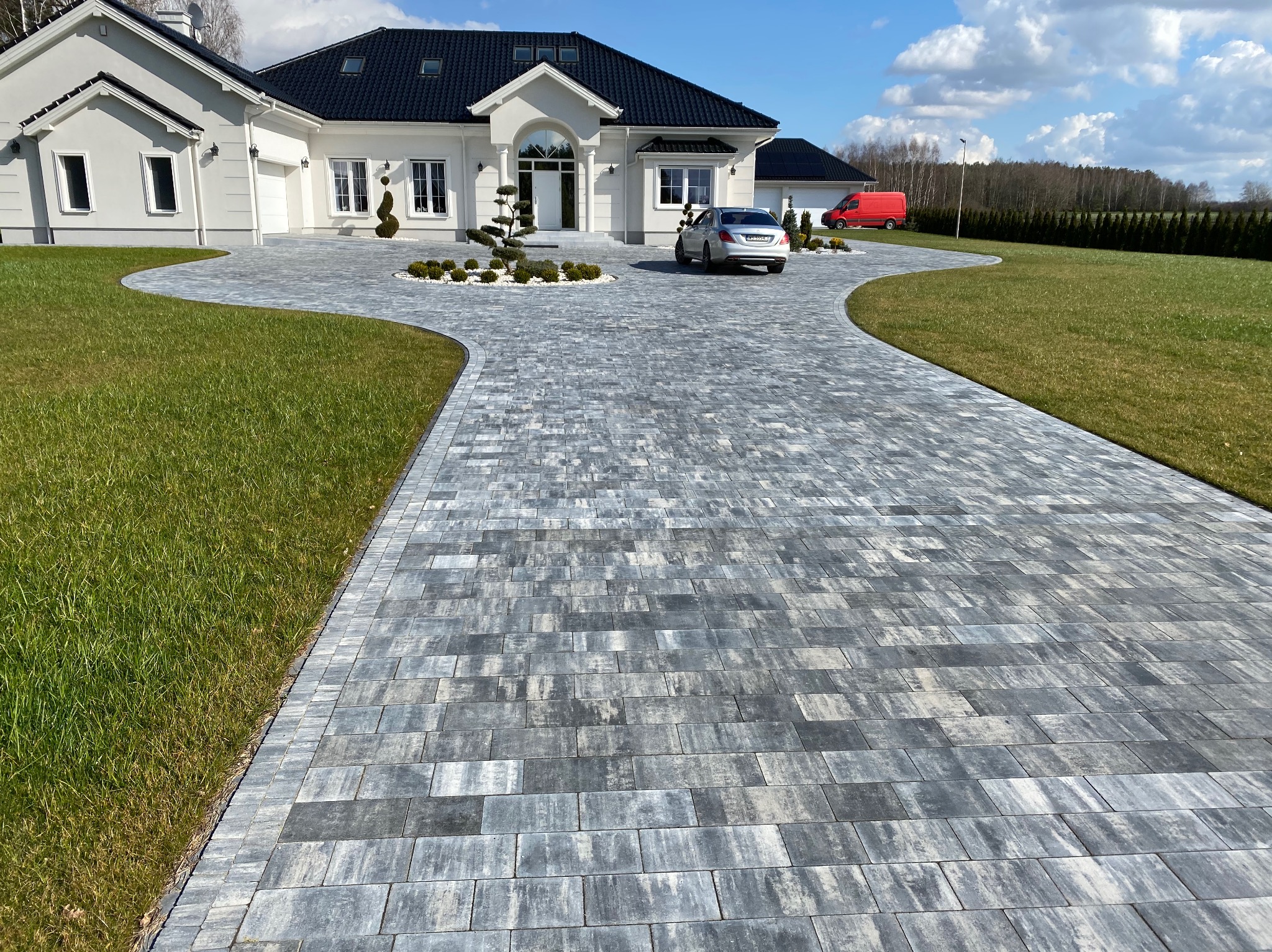 Szeroki, zakrzywiony podjazd z szarej kostki brukowej prowadzący do nowoczesnego, białego domu z czarnym dachem; na podjeździe zaparkowany srebrny samochód, w tle czerwony van i zielony trawnik.