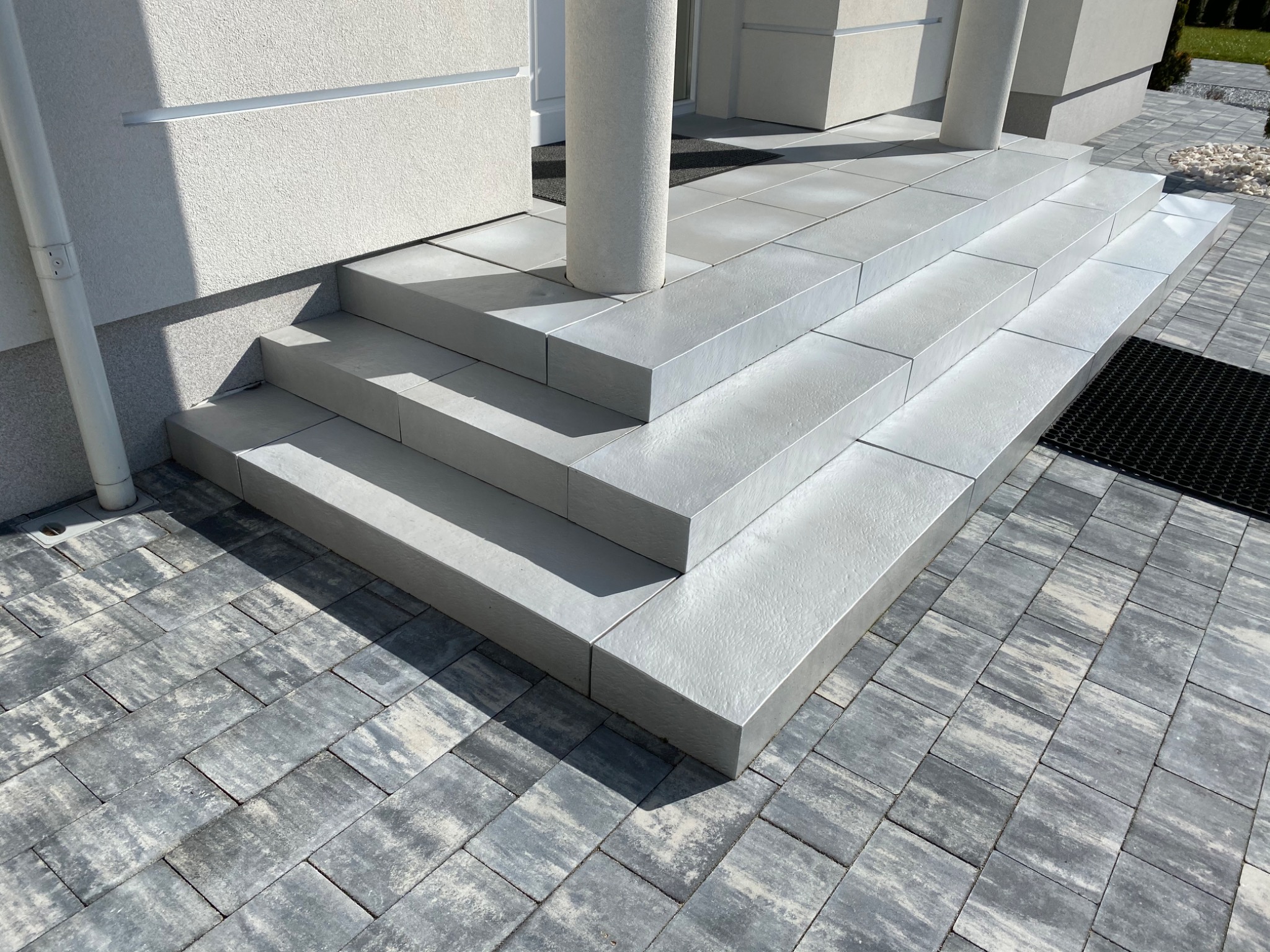 Nowoczesne schody wejściowe z szarej kostki brukowej prowadzące do domu z kolumną, otoczone szarą kostką brukową na podjeździe.