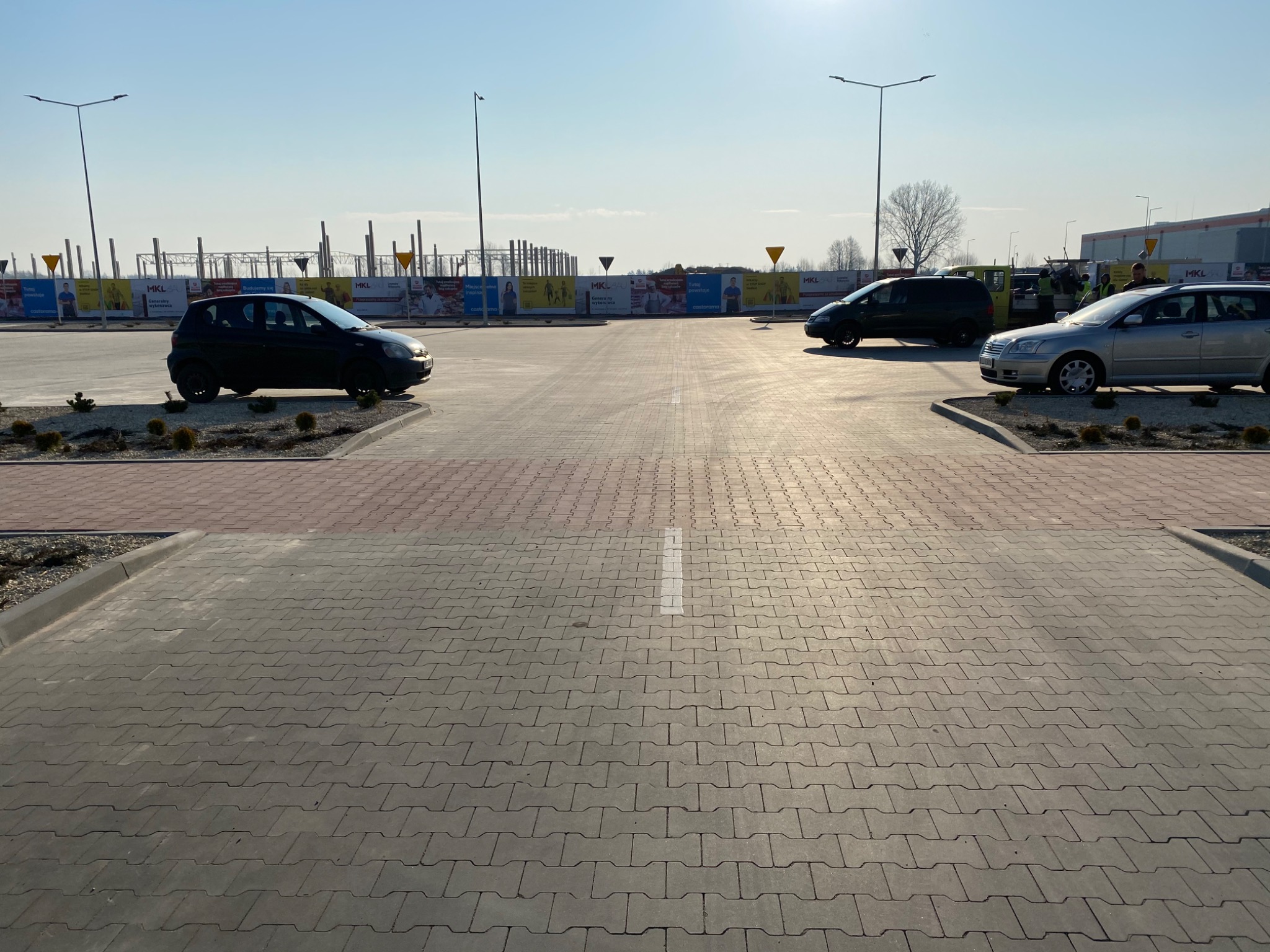 Parking z szarej kostki brukowej w jodełkę, oddzielony pasem czerwonej kostki, z zaparkowanymi samochodami i widocznymi banerami reklamowymi w tle pod błękitnym niebem.