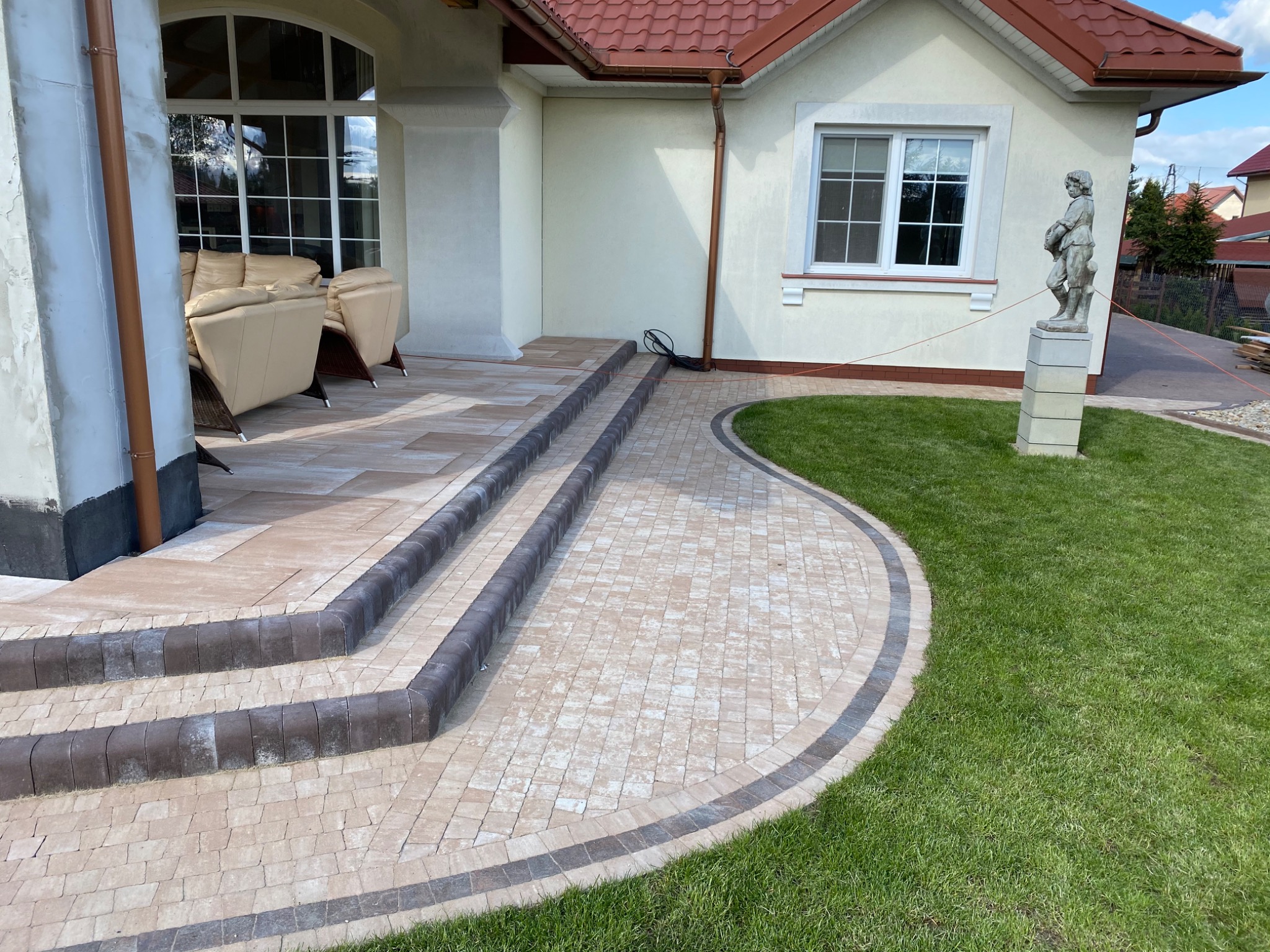 Wykończony podjazd z kostki brukowej w odcieniach beżu i brązu, prowadzący do domu z jasną elewacją, widoczne schody z obrzeżami, trawnik i rzeźba chłopca w tle.