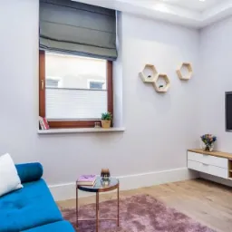 Jasne wnętrze pokoju z błękitną sofą, drewnianym oknem z plisowaną roletą na dole i szarą roletą rzymską na górze, oraz dekoracjami ściennymi w kształcie plastra miodu.