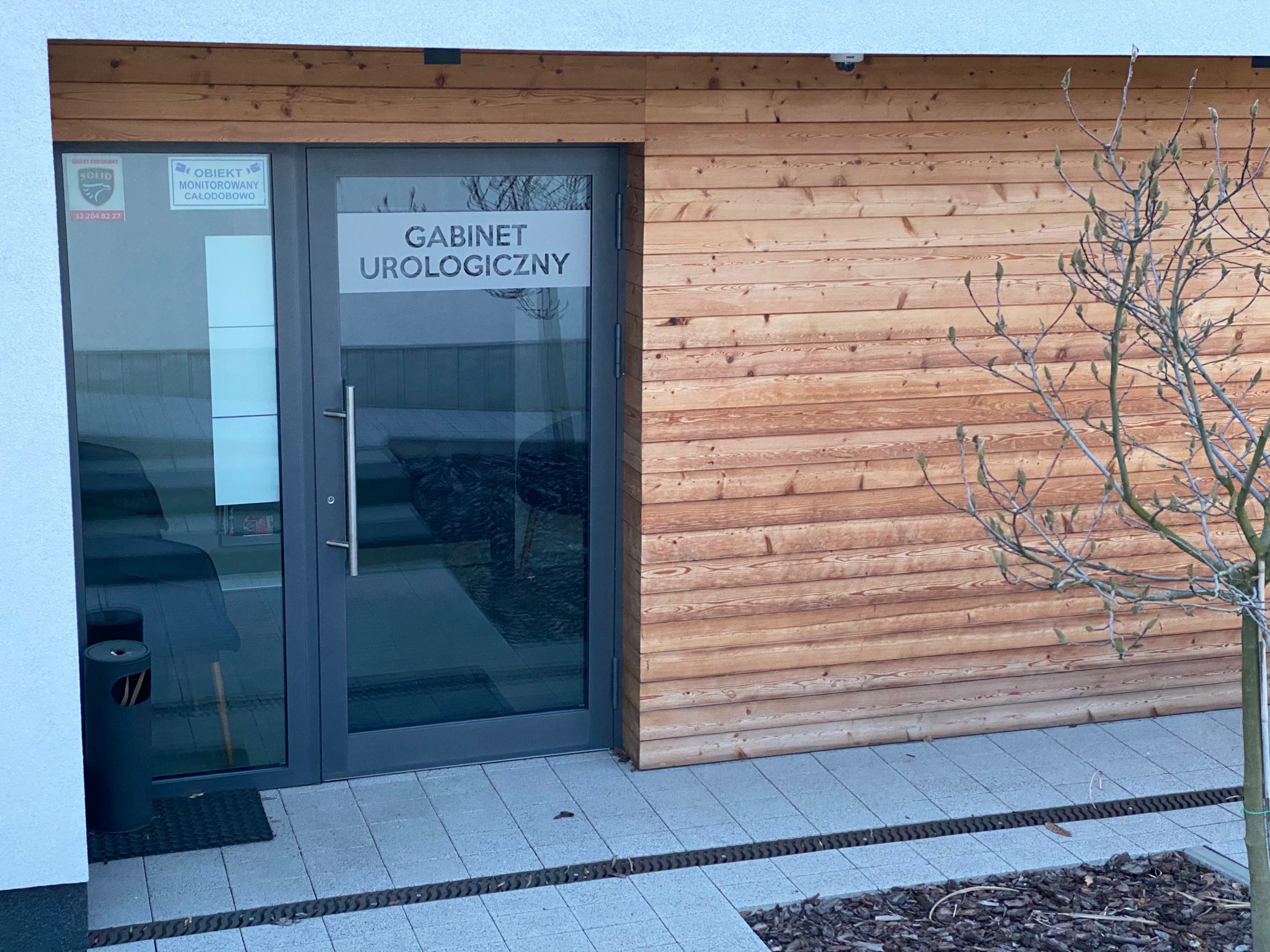 Wejście do gabinetu lekarskiego z napisem 'GABINET UROLOGICZNY' na szklanych drzwiach, obok drewniana elewacja i małe drzewko.