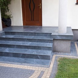 Piotr i Paweł - usługi remontowo wykończeniowe - Zewnętrzne schody wejściowe wyłożone szarą płytką imitującą kamień, z widocznym fragmentem elewacji, drewnianych drzwi wejściowych i kostki brukowej w odcieniach szarości i beżu.