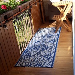 Piotr i Paweł - usługi remontowo wykończeniowe - Wykończony balkon z drewnianą podłogą, niebiesko-białym dywanem w orientalne wzory, drewnianymi meblami ogrodowymi i kwiatami w donicach na balustradzie.