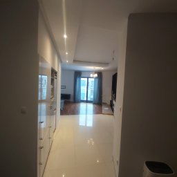 Piotr i Paweł - usługi remontowo wykończeniowe - Nowoczesne wnętrze mieszkania z białymi, błyszczącymi płytkami podłogowymi, zabudową kuchenną na lewo i widokiem na salon z balkonem w tle.