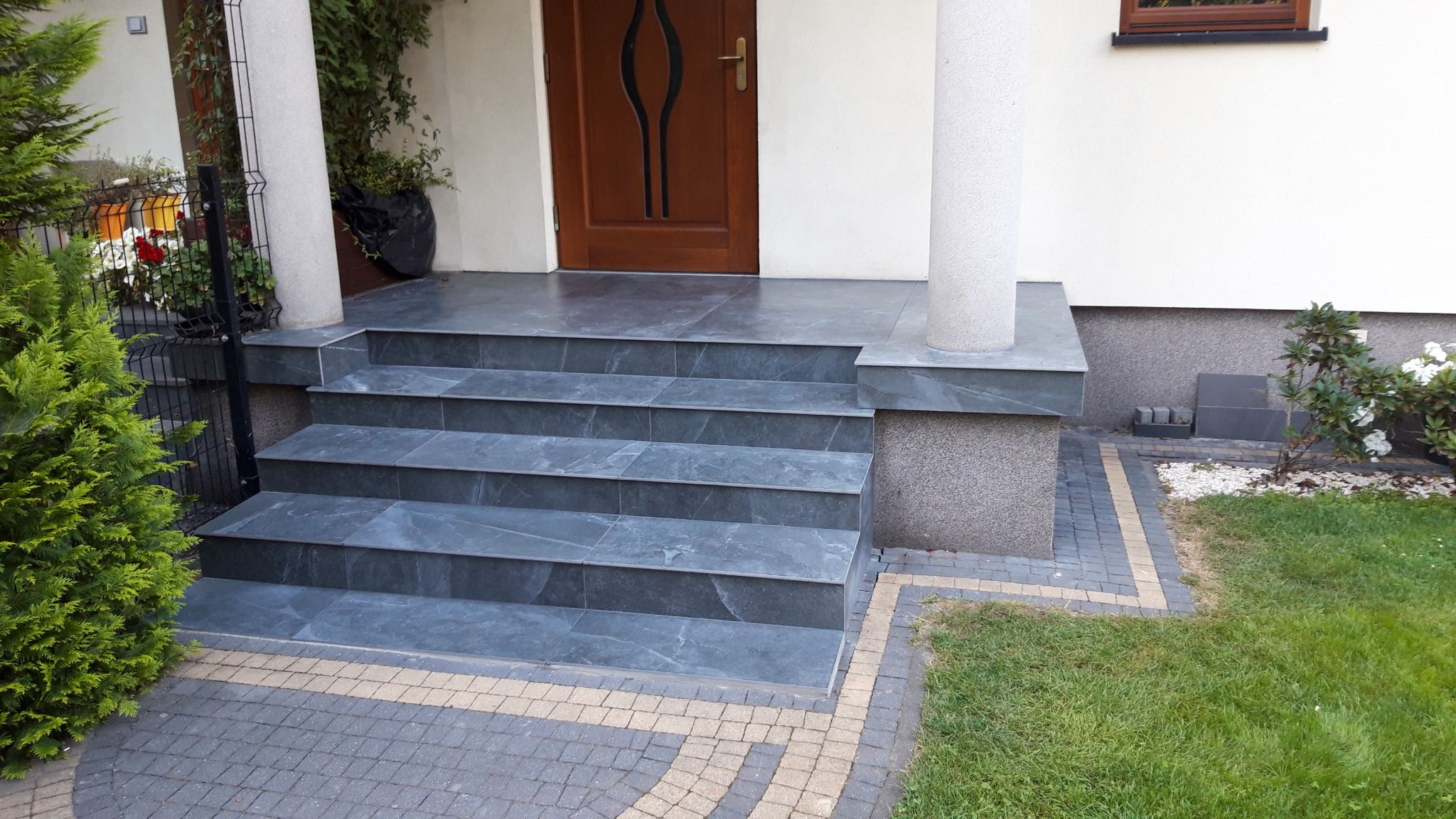 Zewnętrzne schody wejściowe wyłożone szarą płytką imitującą kamień, z widocznym fragmentem elewacji, drewnianych drzwi wejściowych i kostki brukowej w odcieniach szarości i beżu.