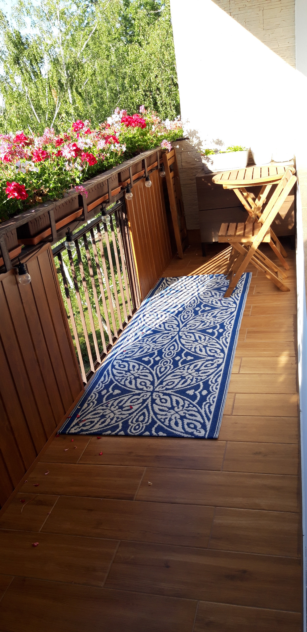 Wykończony balkon z drewnianą podłogą, niebiesko-białym dywanem w orientalne wzory, drewnianymi meblami ogrodowymi i kwiatami w donicach na balustradzie.