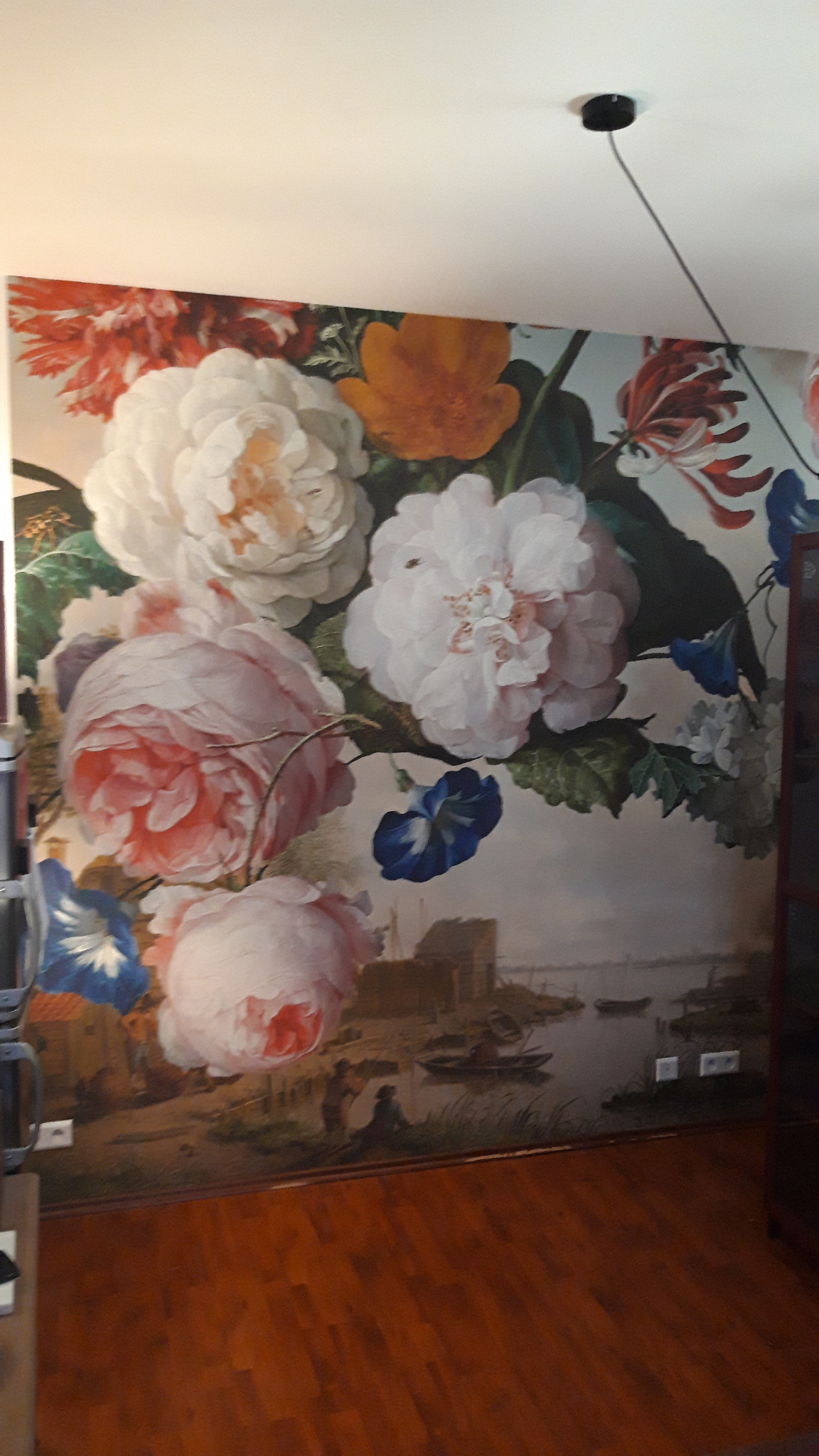 Ściana z fototapetą przedstawiającą bukiet kwiatów w stylu holenderskim na tle pejzażu z łodziami, uzupełniona drewnianą podłogą i czarnym punktowym oświetleniem sufitowym.