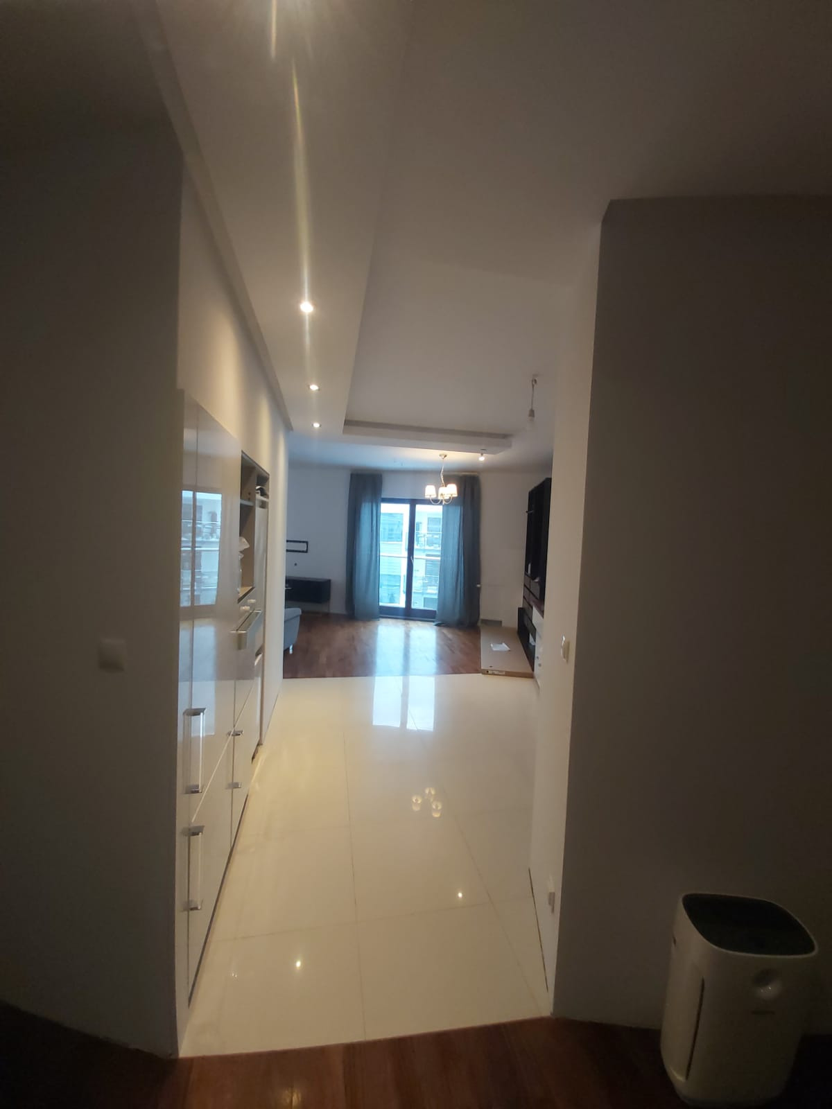 Nowoczesne wnętrze mieszkania z białymi, błyszczącymi płytkami podłogowymi, zabudową kuchenną na lewo i widokiem na salon z balkonem w tle.