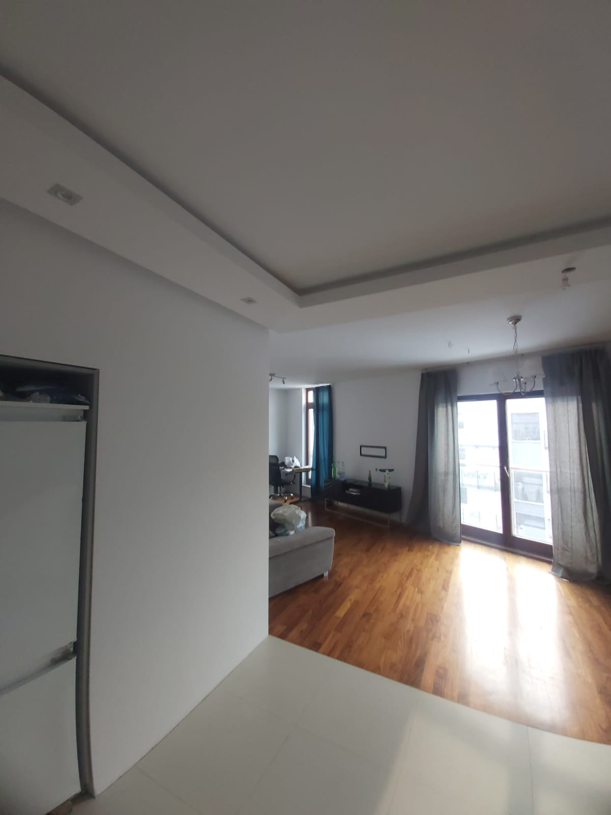 Nowoczesne wnętrze po remoncie: białe ściany, parkiet, sufit z oświetleniem punktowym i widok na balkon przez przesuwne drzwi.