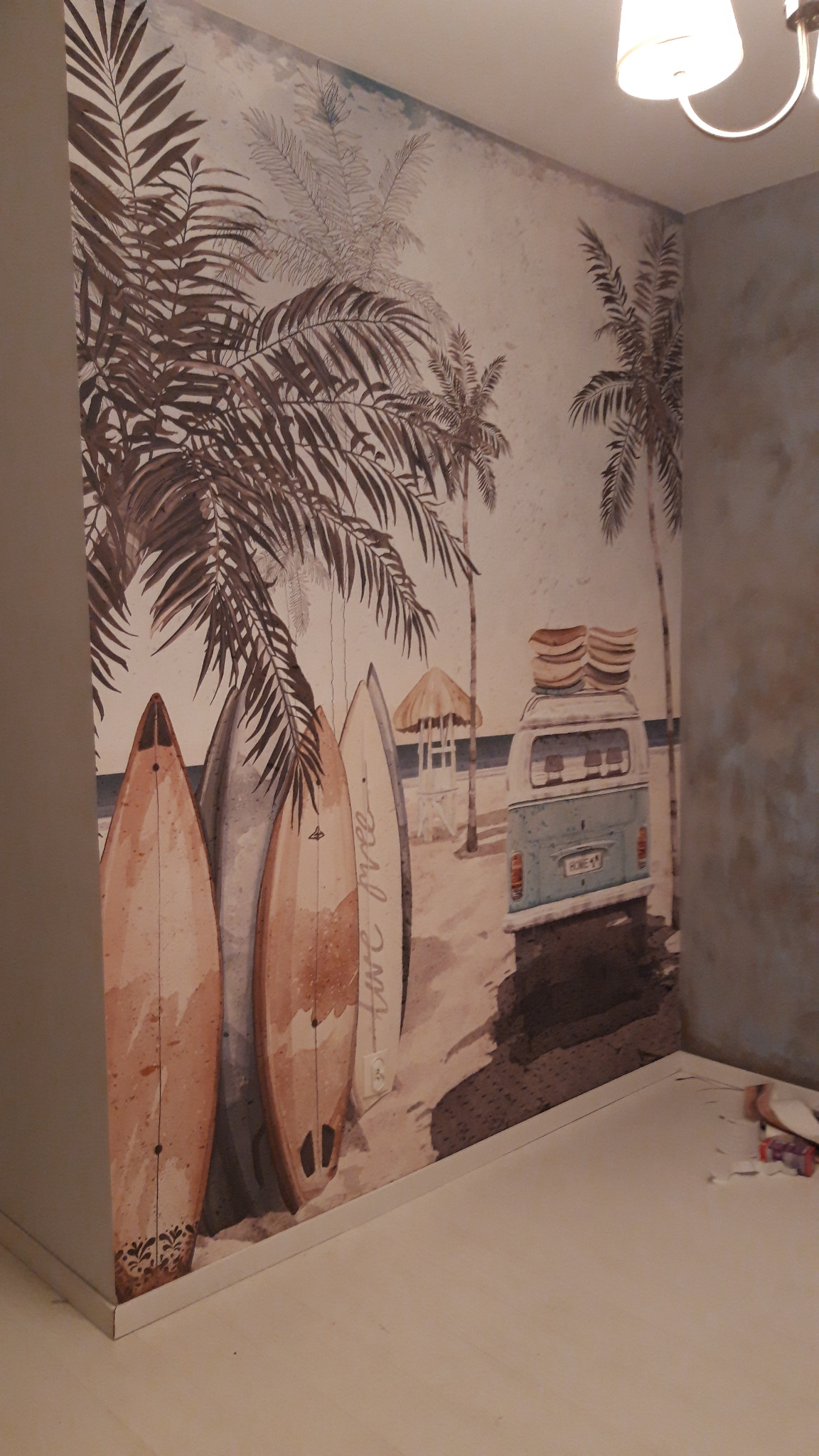 Ściana z fototapetą przedstawiającą plażę z palmami, deskami surfingowymi i busem z deskami na dachu, widoczne niedoskonałości w narożniku i przy podłodze.