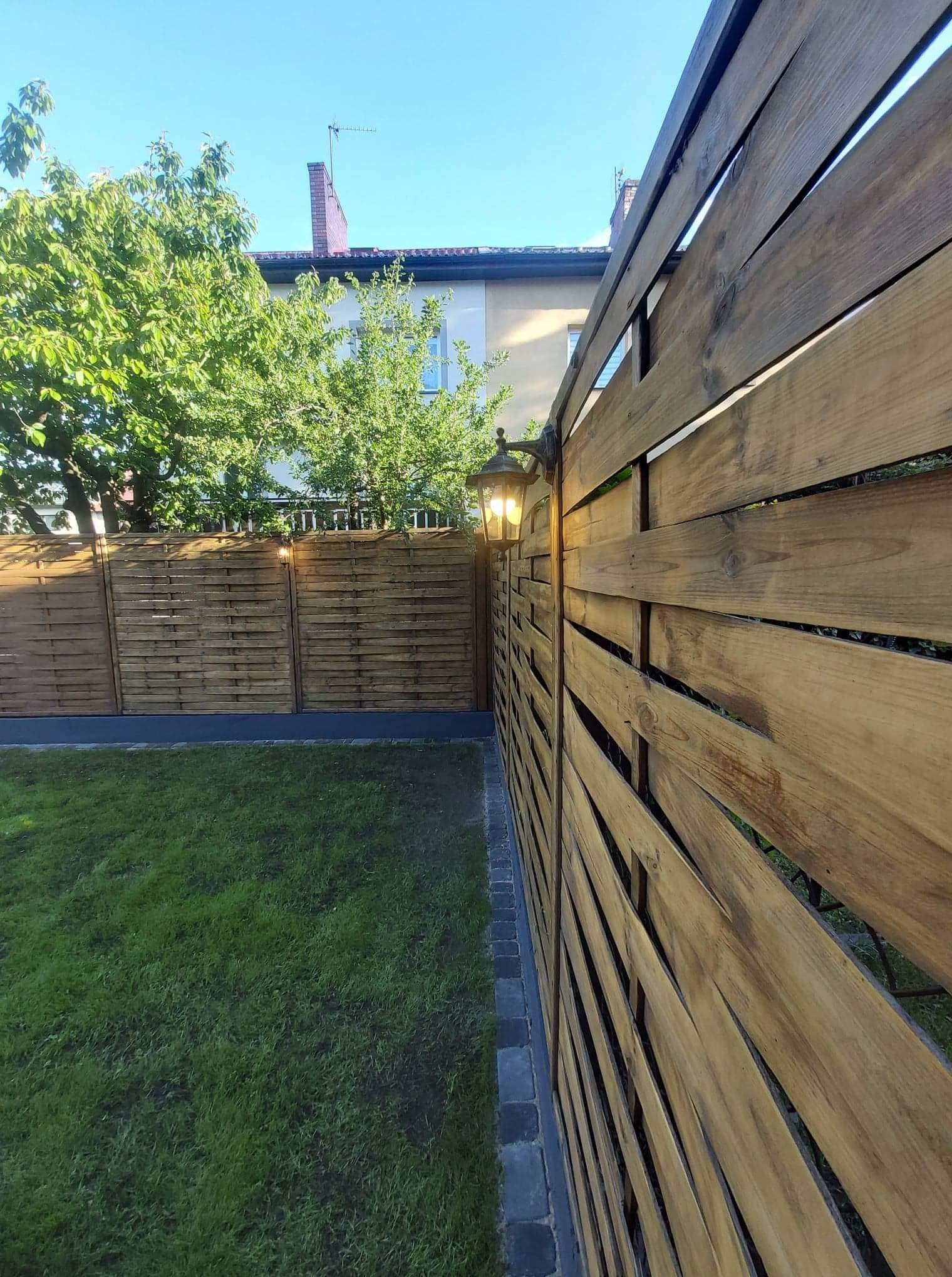 Nowoczesne drewniane ogrodzenie panelowe z oświetleniem zewnętrznym, widok na trawnik i budynek w tle w słoneczny dzień.