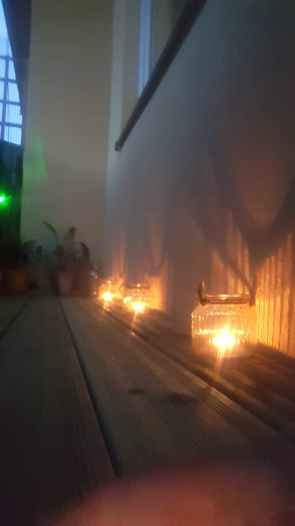 Rząd szklanych lampionów ze świecami ustawionych na drewnianym parapecie wzdłuż ściany, tworzących ciepłe, rozproszone światło.