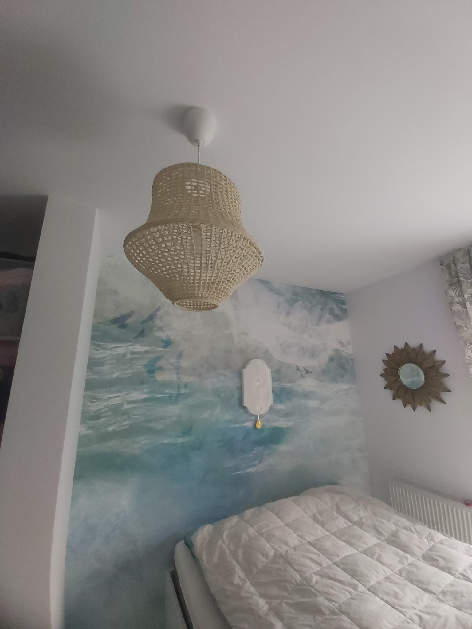 Sypialnia z tapetą imitującą morski krajobraz, wiklinowa lampa wisząca i lustro w kształcie słońca na ścianie.