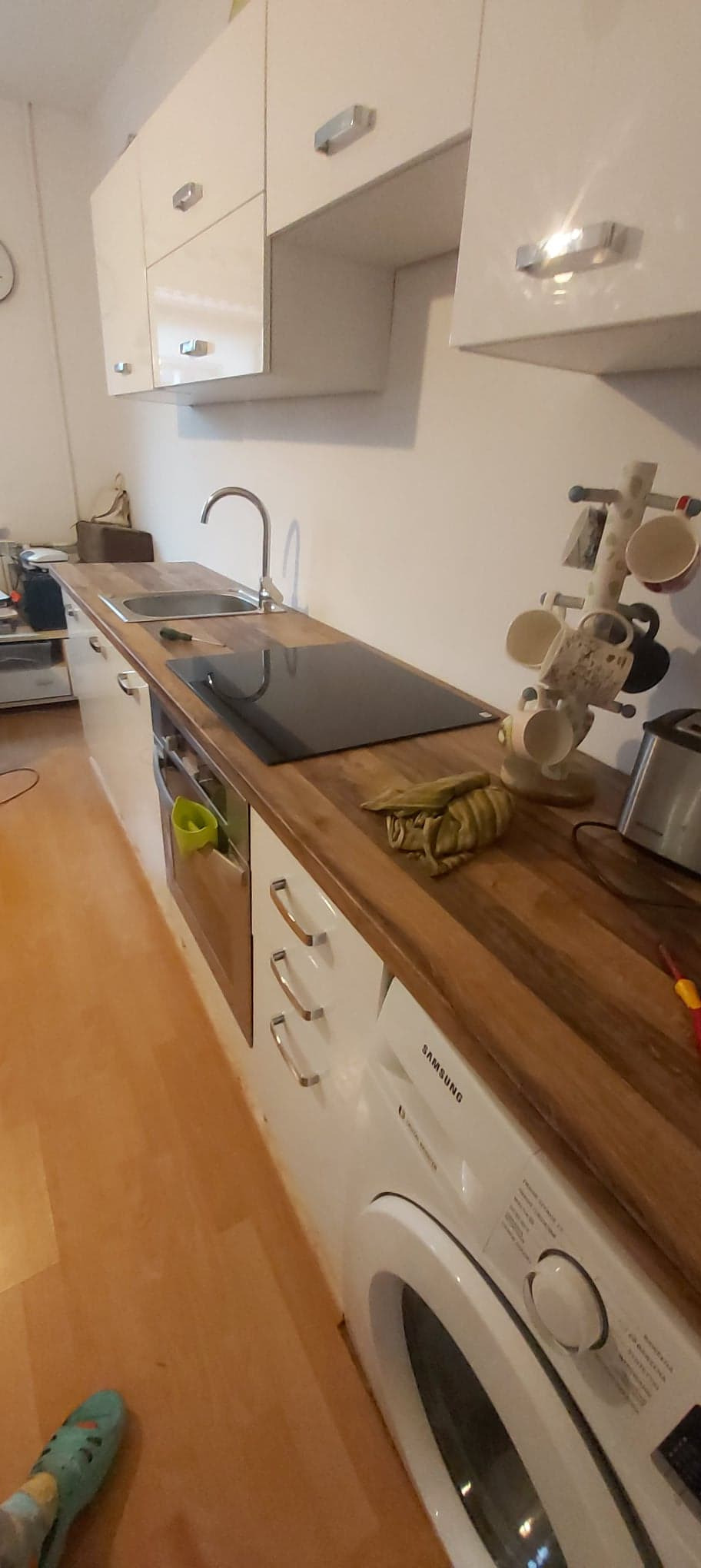 Wnętrze kuchni z białymi szafkami, drewnianym blatem, czarną płytą indukcyjną, zlewozmywakiem i stojakiem na kubki, z pralką pod blatem