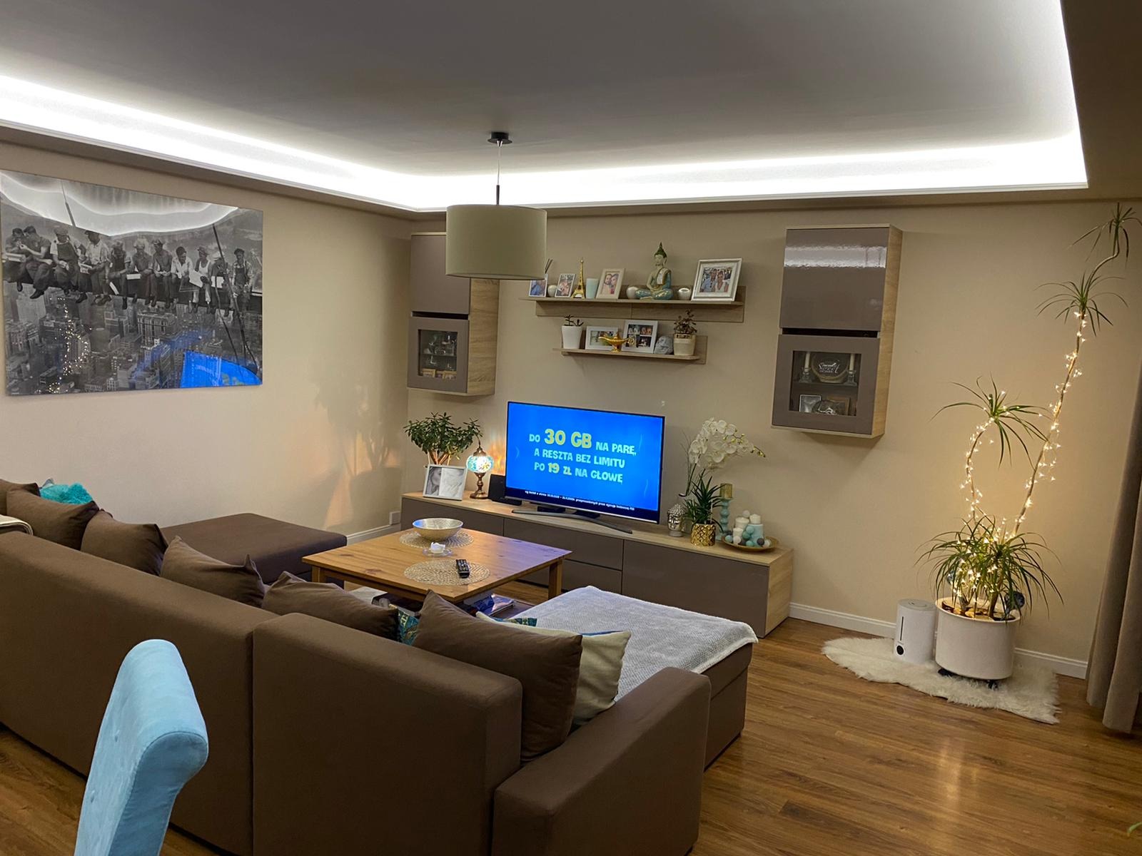 Jasne wnętrze salonu z brązową sofą narożną, drewnianą podłogą i sufitem z oświetleniem LED, na ścianie obraz z motywem budowlanym, telewizor i wiszące szafki.