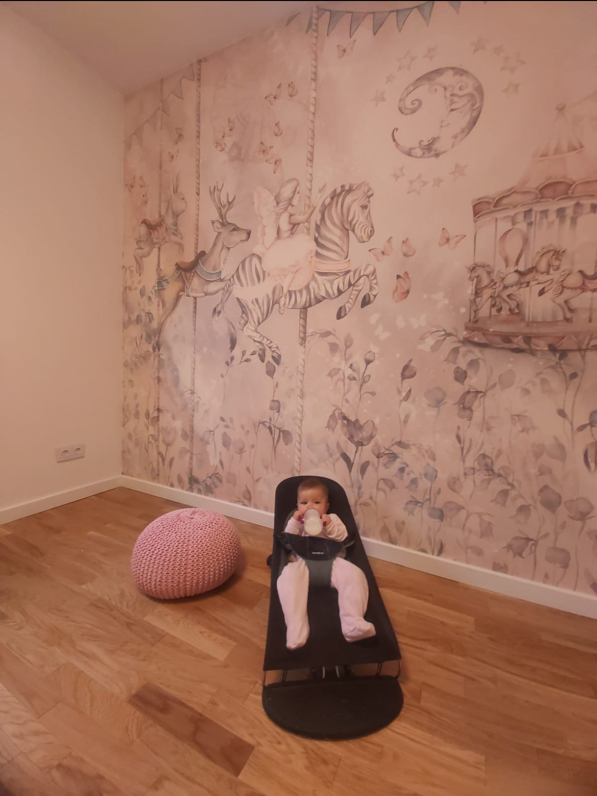 Pokój dziecięcy w Karczewie z tapetą przedstawiającą motywy karuzeli, w foteliku siedzi dziecko pijące mleko, obok pufa.