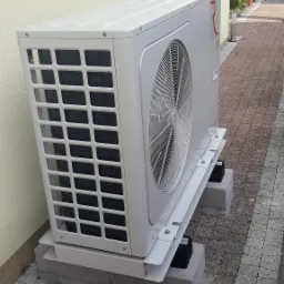 Zewnętrzna jednostka klimatyzacyjna Rotenso zamontowana na betonowych podkładach przy ścianie budynku z beżowym tynkiem, widoczne ogrodzenie posesji w tle.