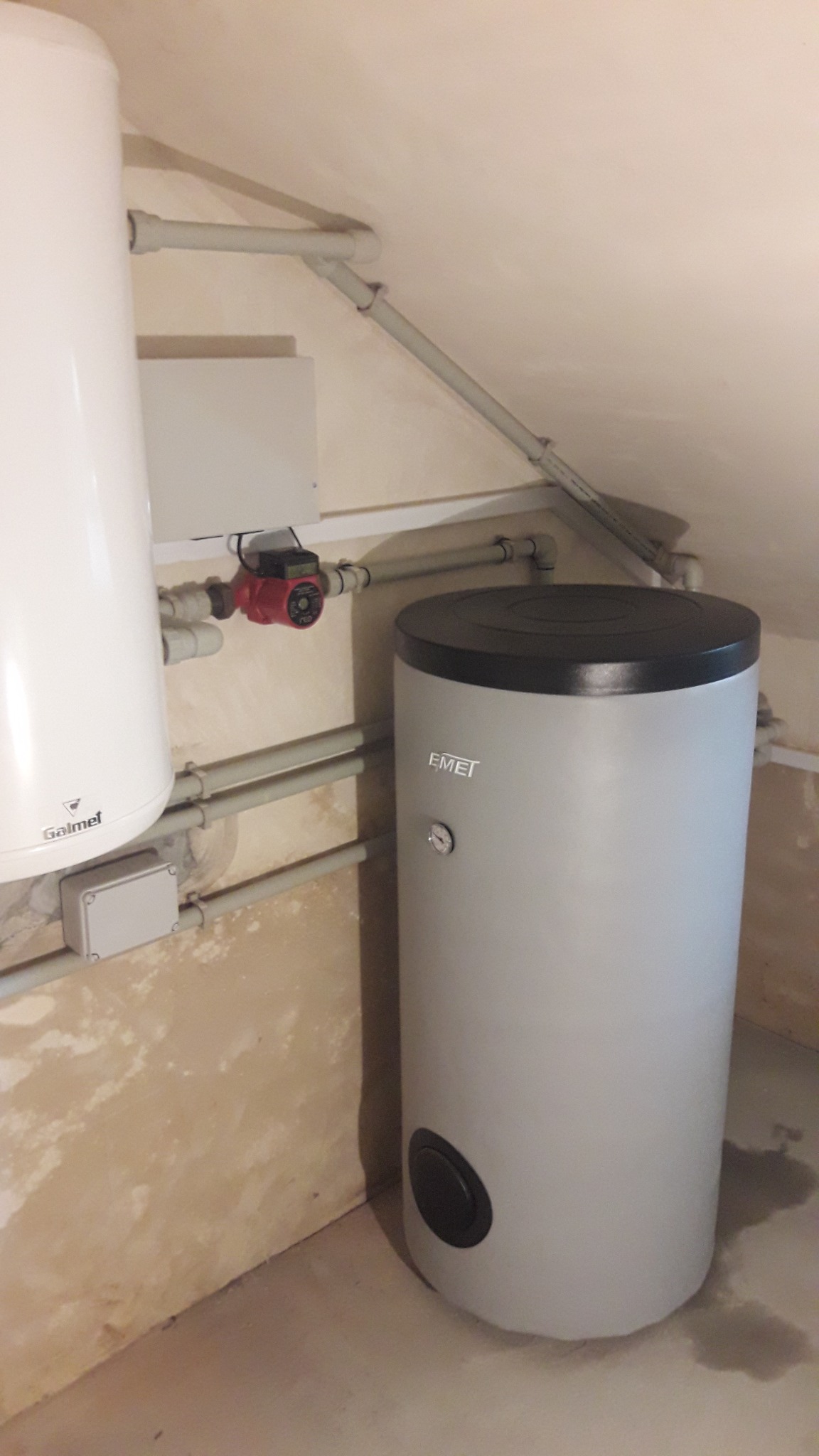 Dwa zasobniki ciepłej wody użytkowej, jeden marki Galmet, drugi Emet, połączone rurami z czerwoną pompą obiegową, zamontowane w pomieszczeniu z surowymi ścianami.