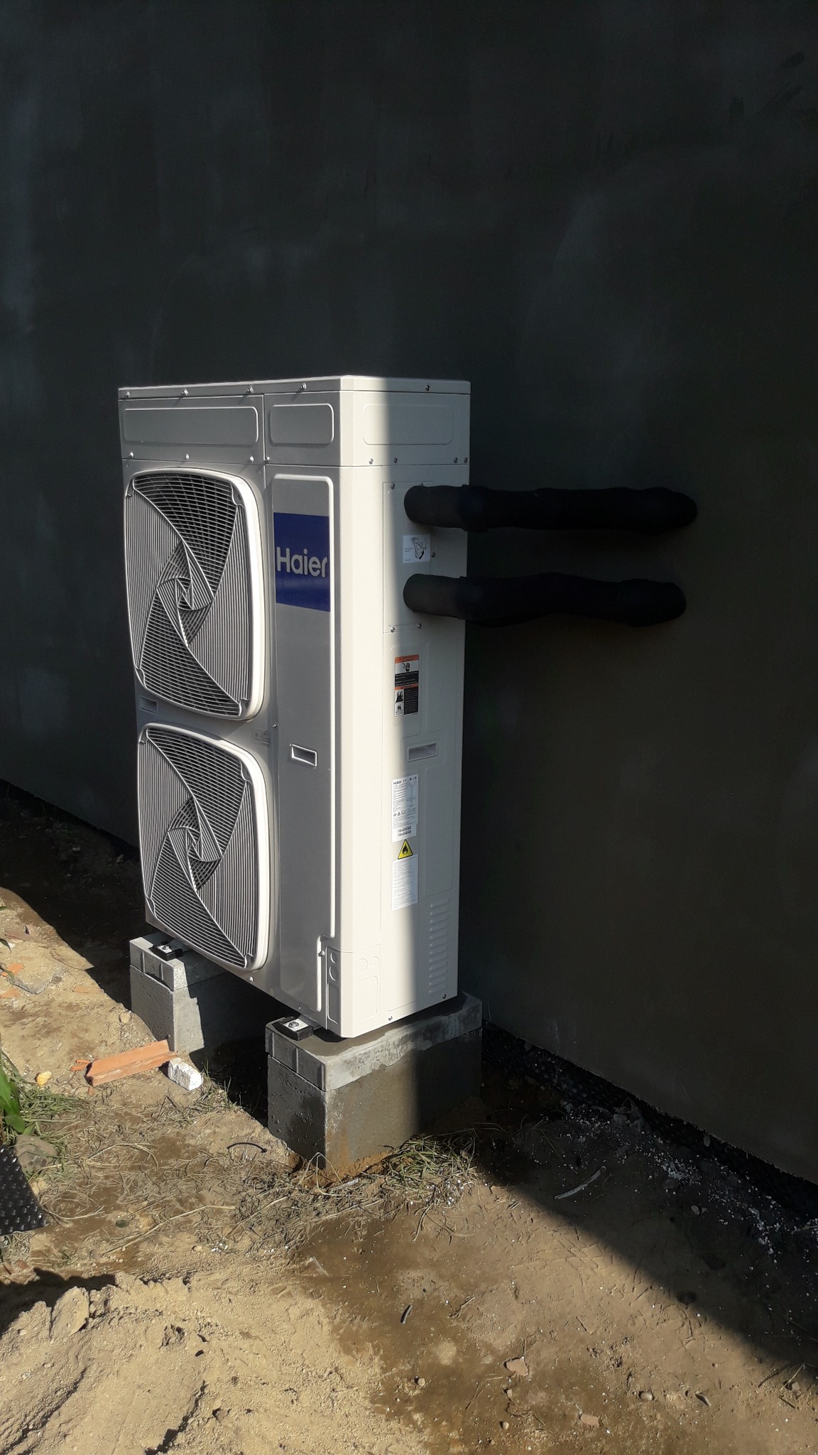 Zewnętrzna jednostka klimatyzacji Haier zamontowana na betonowych bloczkach przy szarej ścianie, z widocznymi izolowanymi rurami chłodniczymi.