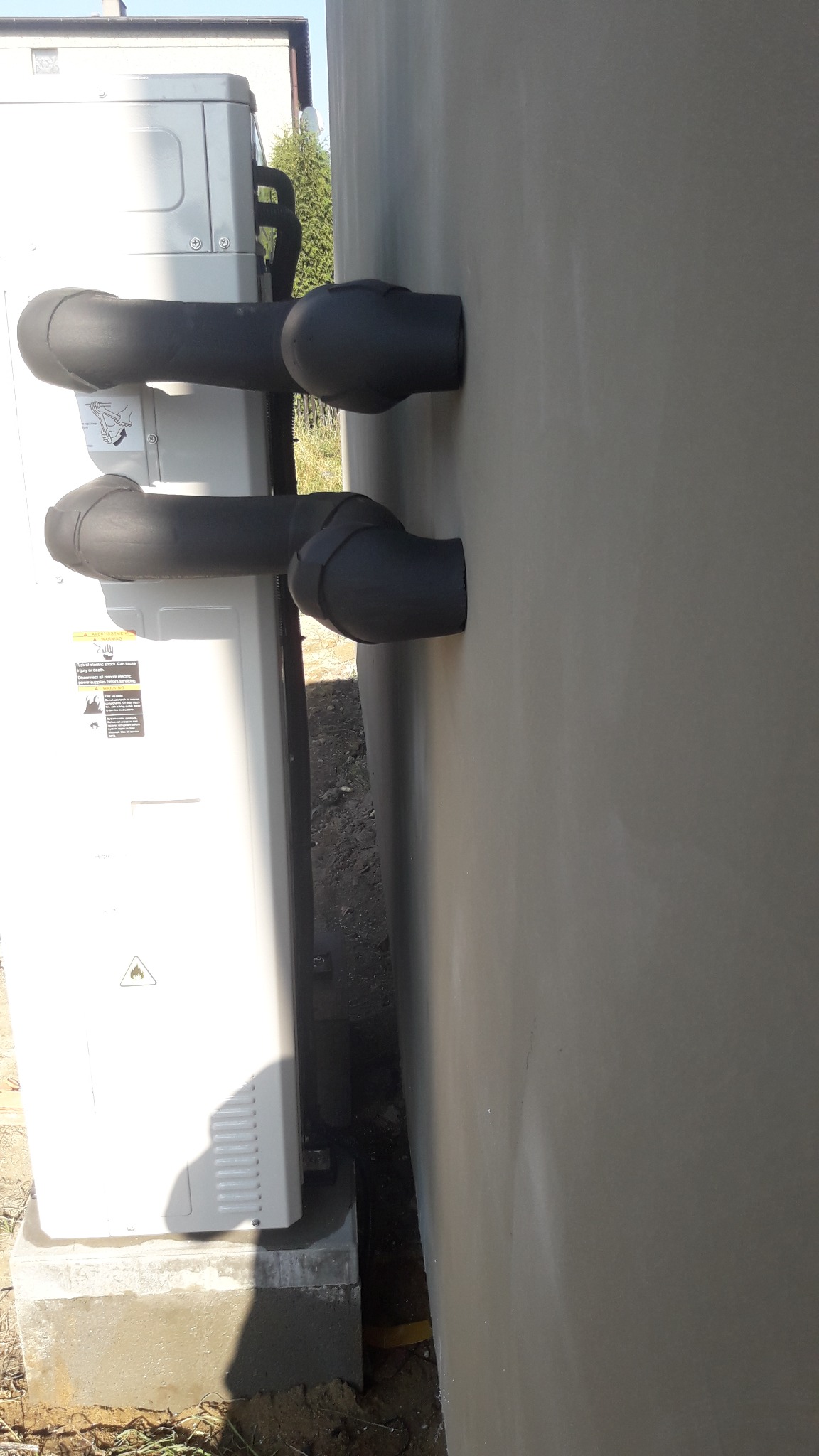 Zewnętrzna jednostka klimatyzacyjna z podłączonymi, zaizolowanymi rurami wchodzącymi do budynku przez ścianę, zamontowana na betonowej podstawie.